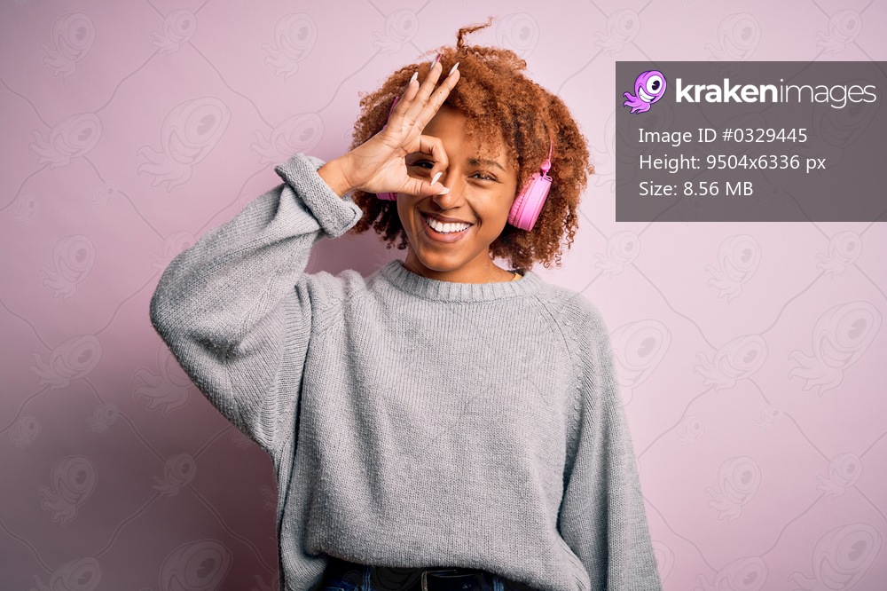 Young African American afro woman with curly hair listening to music using pink headphones doing ok gesture with hand smiling, eye looking through fingers with happy face.