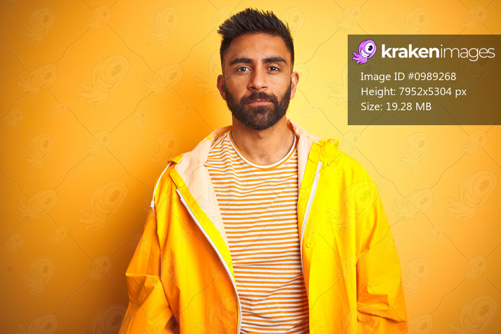 Young indian man wearing raincoat standing over isolated yellow background Relaxed with serious expression on face. Simple and natural looking at the camera.