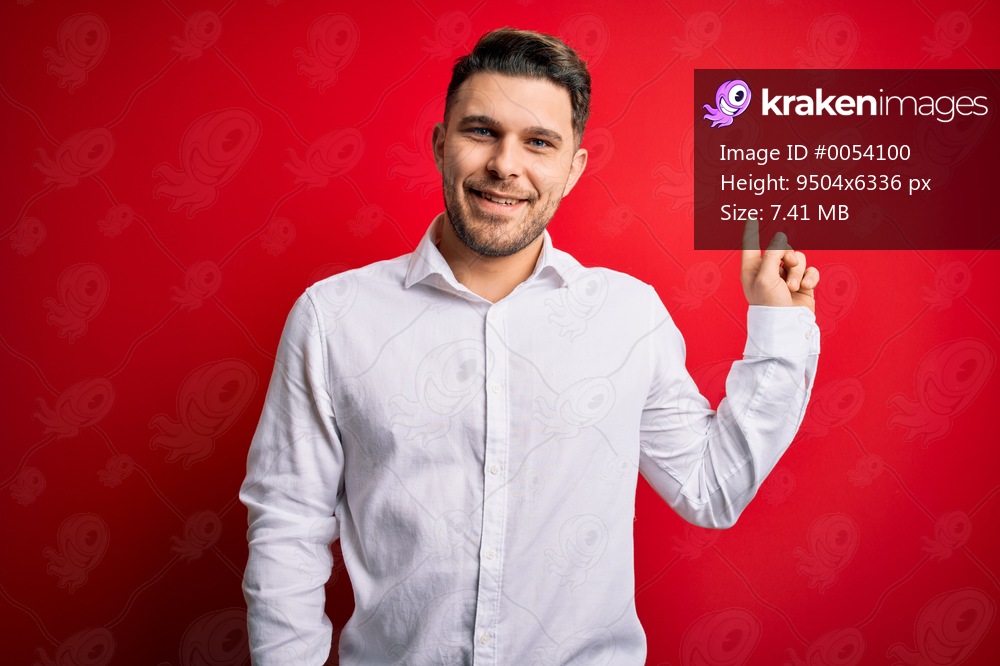 Young business man with blue eyes wearing elegant shirt standing over red isolated background with a big smile on face, pointing with hand finger to the side looking at the camera.