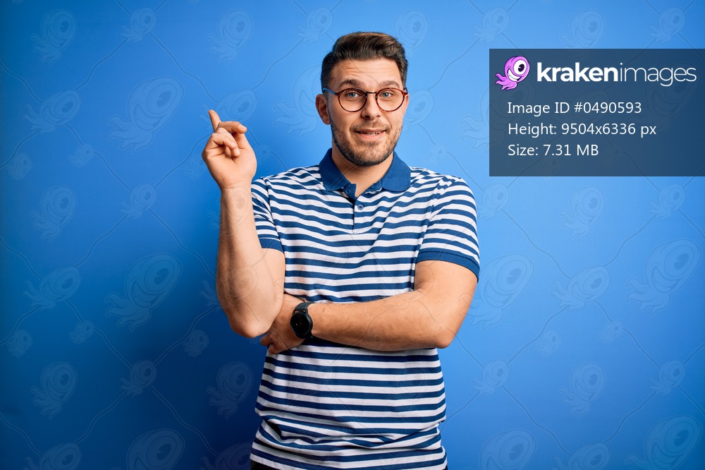 Young man with blue eyes wearing glasses and casual striped t-shirt over blue background with a big smile on face, pointing with hand and finger to the side looking at the camera.
