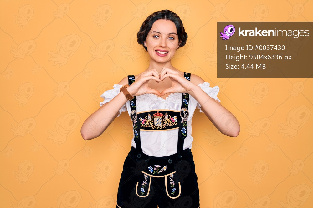 Young beautiful german woman with blue eyes wearing traditional octoberfest dress smiling in love doing heart symbol shape with hands. Romantic concept.
