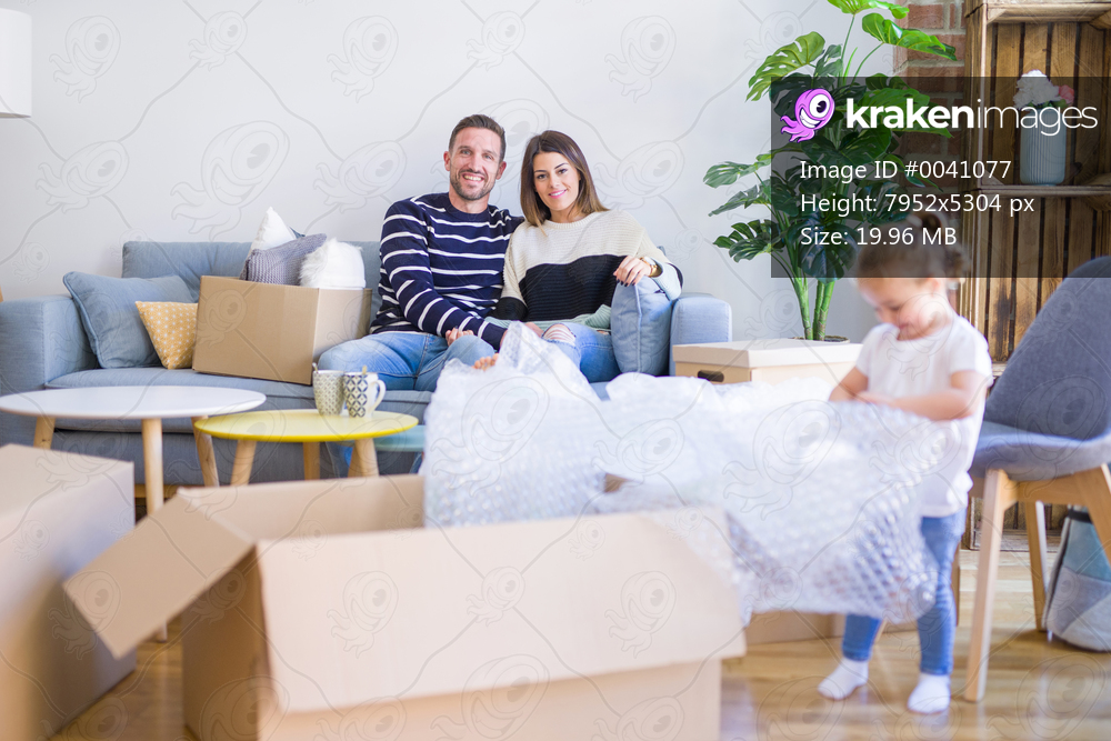 Beautiful family, parents sitting on the sofa drinking coffee looking his kid playing at new home around cardboard boxes
