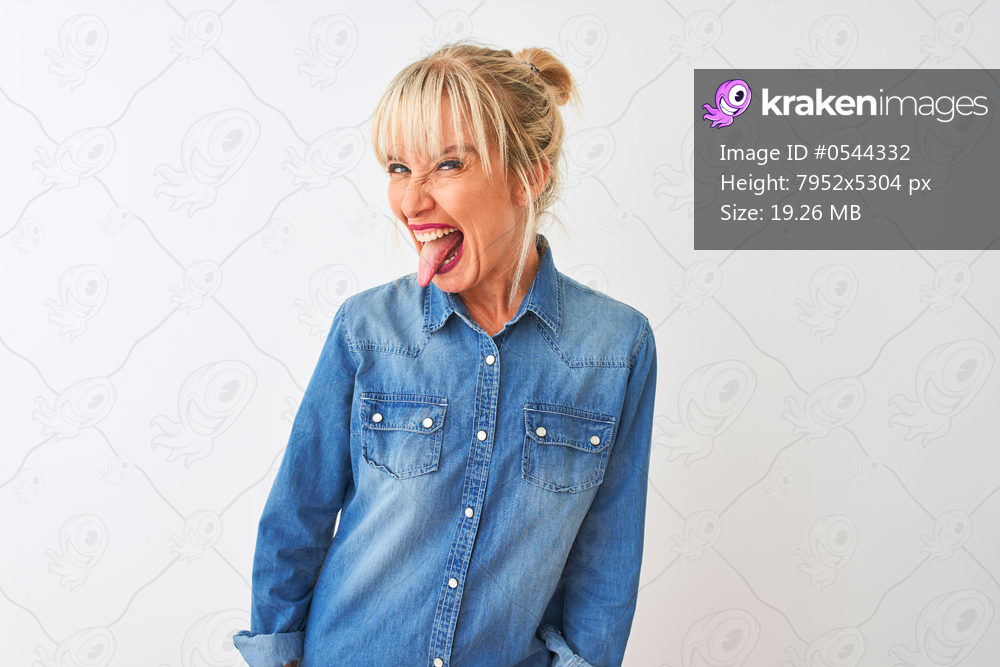 Middle age woman wearing casual denim shirt standing over isolated white background sticking tongue out happy with funny expression. Emotion concept.