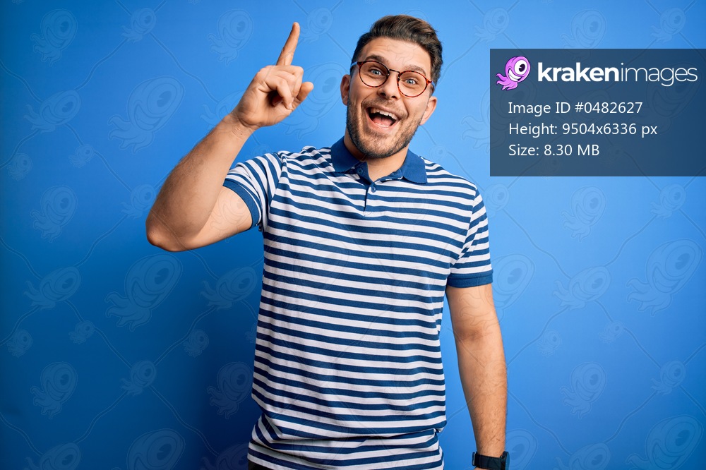 Young man with blue eyes wearing glasses and casual striped t-shirt over blue background pointing finger up with successful idea. Exited and happy. Number one.