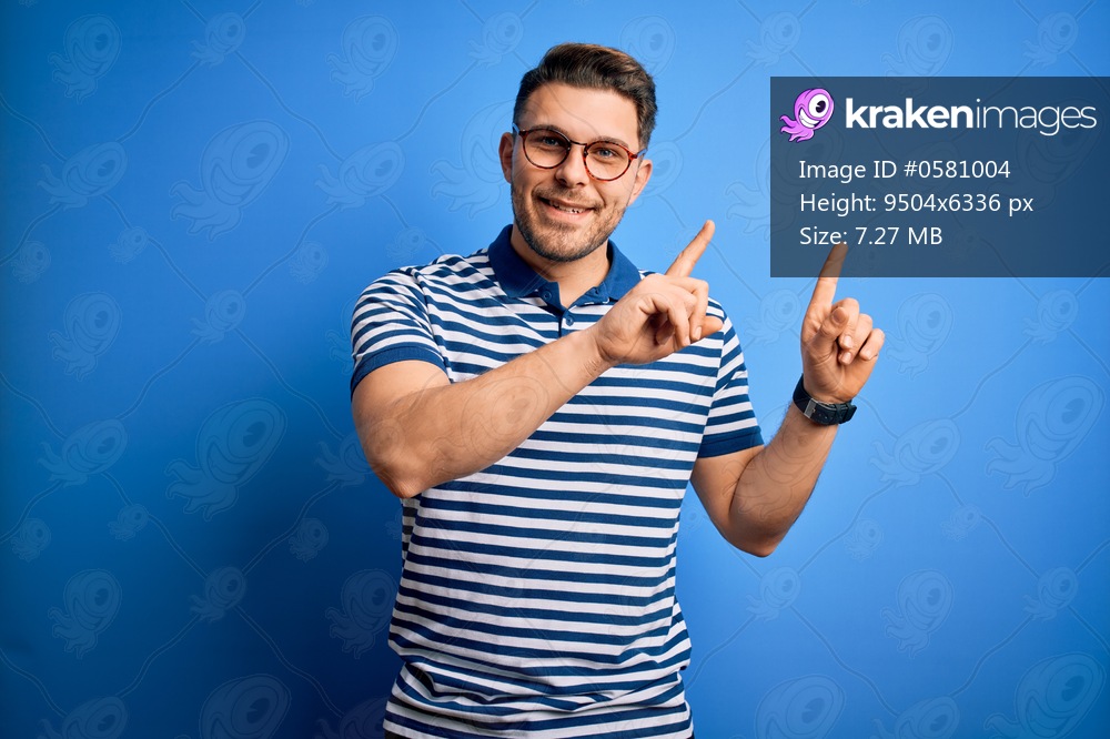 Young man with blue eyes wearing glasses and casual striped t-shirt over blue background smiling and looking at the camera pointing with two hands and fingers to the side.