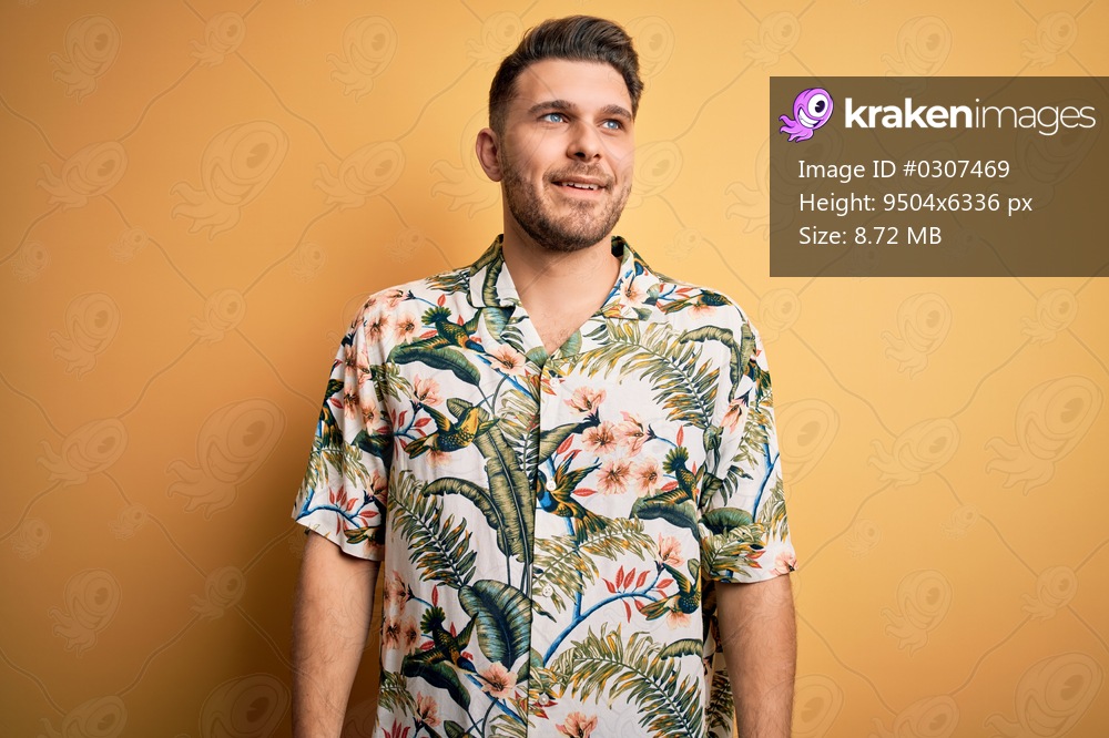 Young man with blue eyes on vacation wearing floral summer shirt over yellow background looking away to side with smile on face, natural expression. Laughing confident.