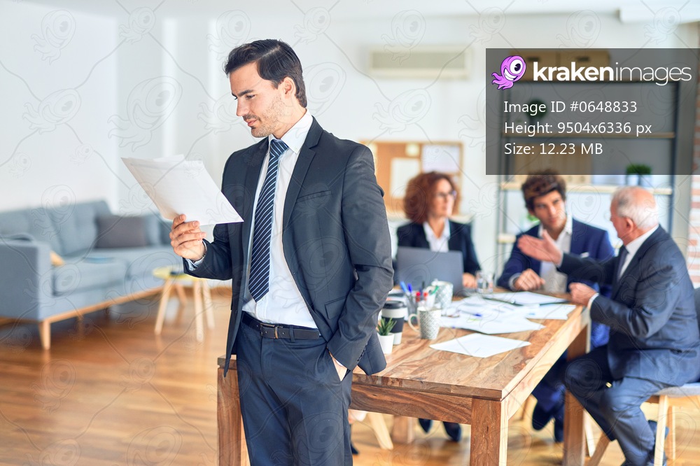 Group of business workers smiling happy and confident working together in a meeting. One of them, standing with smile on face reading documents at the office.