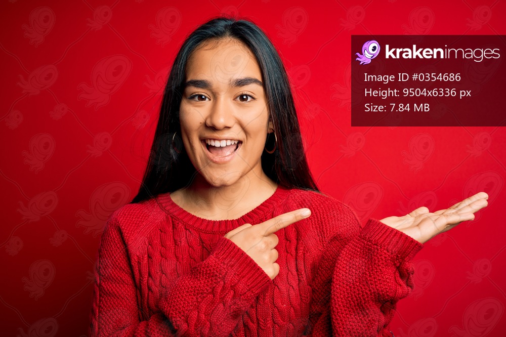 Young beautiful asian woman wearing casual sweater standing over isolated red background amazed and smiling to the camera while presenting with hand and pointing with finger.