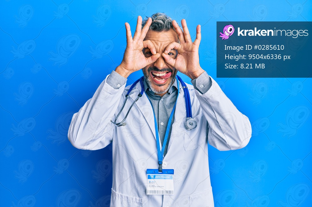 Middle age grey-haired man wearing doctor uniform and stethoscope doing ok gesture like binoculars sticking tongue out, eyes looking through fingers. crazy expression. 