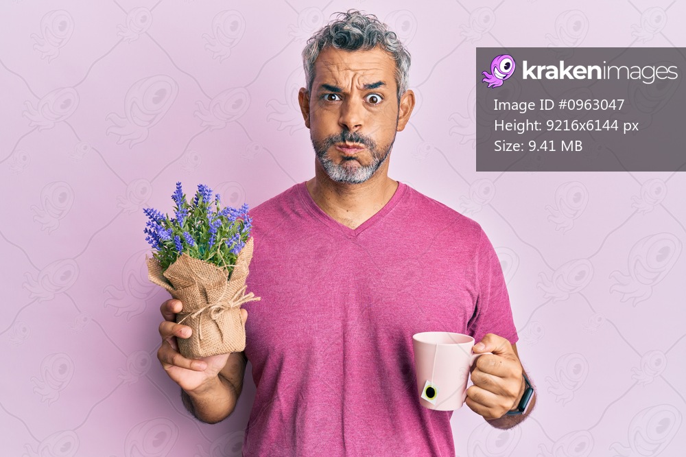 Middle age grey-haired man drinking a cup of infused lavender puffing cheeks with funny face. mouth inflated with air, catching air. 