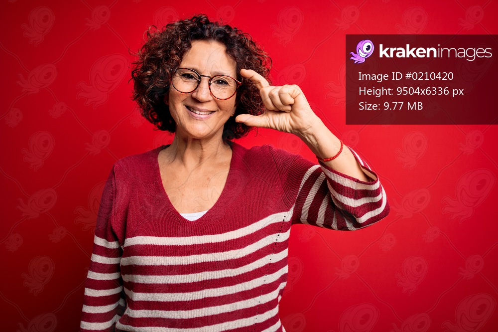 Middle age beautiful curly hair woman wearing casual striped sweater over red background smiling and confident gesturing with hand doing small size sign with fingers looking and the camera. Measure concept.