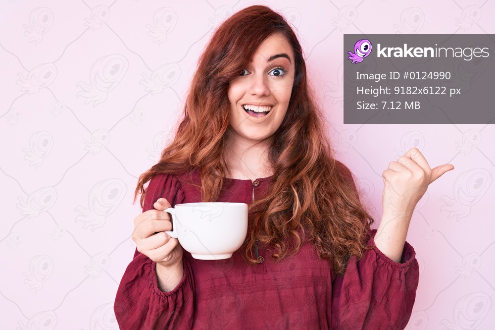Young beautiful woman holding coffee pointing thumb up to the side smiling happy with open mouth 