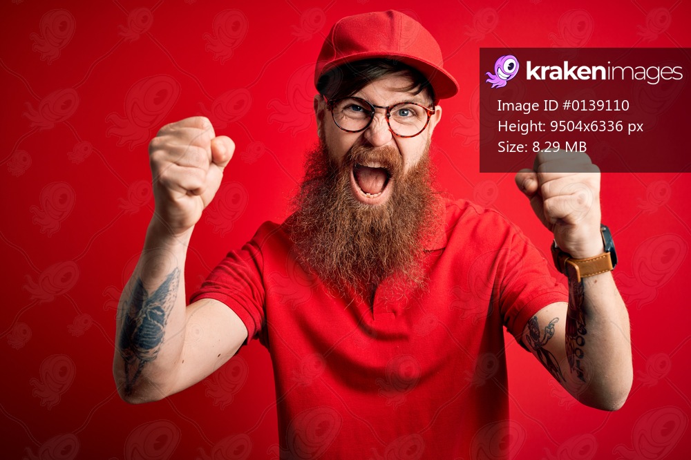 Young handsome delivery man wearing glasses and red cap over isolated background angry and mad raising fists frustrated and furious while shouting with anger. Rage and aggressive concept.