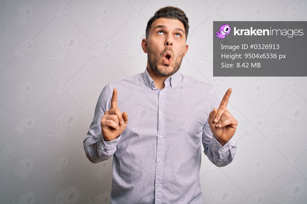 Young business man with blue eyes standing over isolated background amazed and surprised looking up and pointing with fingers and raised arms.