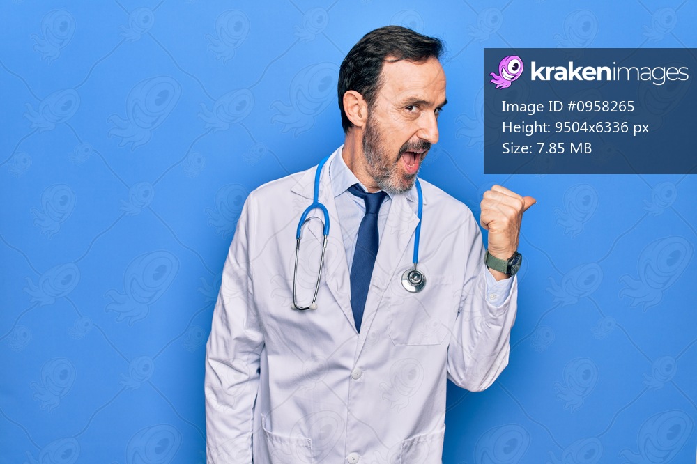 Middle age handsome doctor man wearing coat and stethoscope over isolated blue background pointing thumb up to the side smiling happy with open mouth