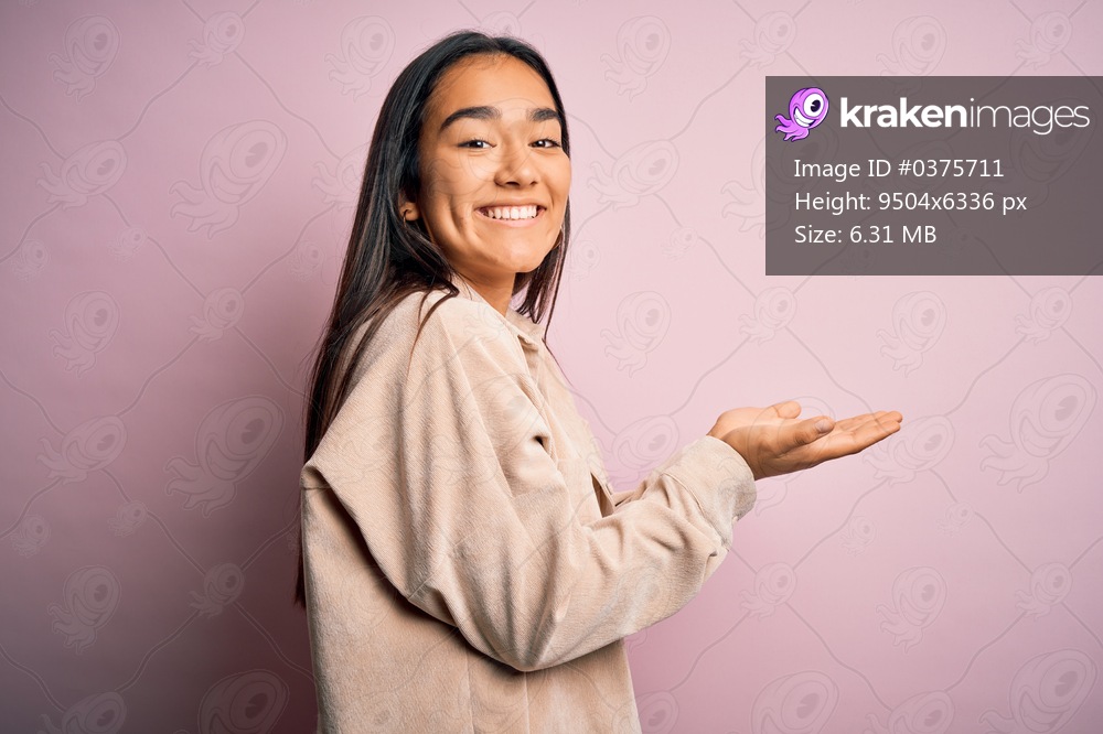 Young beautiful asian woman wearing casual shirt standing over pink background pointing aside with hands open palms showing copy space, presenting advertisement smiling excited happy
