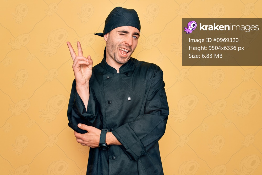Young handsome cooker man with blue eyes wearing uniform and hat over yellow background smiling with happy face winking at the camera doing victory sign. Number two.