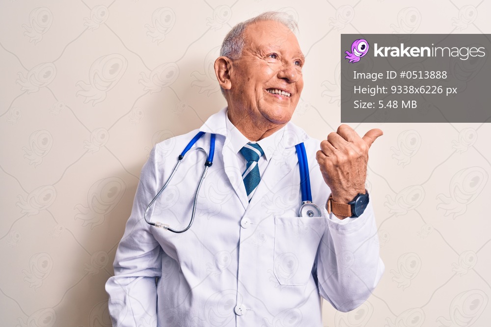Senior grey-haired doctor man wearing coat and stethoscope standing over white background pointing thumb up to the side smiling happy with open mouth