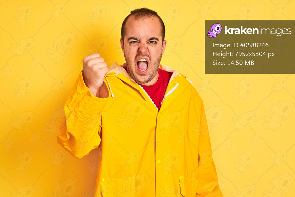 Young man wearing rain coat standing over isolated yellow background angry and mad raising fist frustrated and furious while shouting with anger. Rage and aggressive concept.
