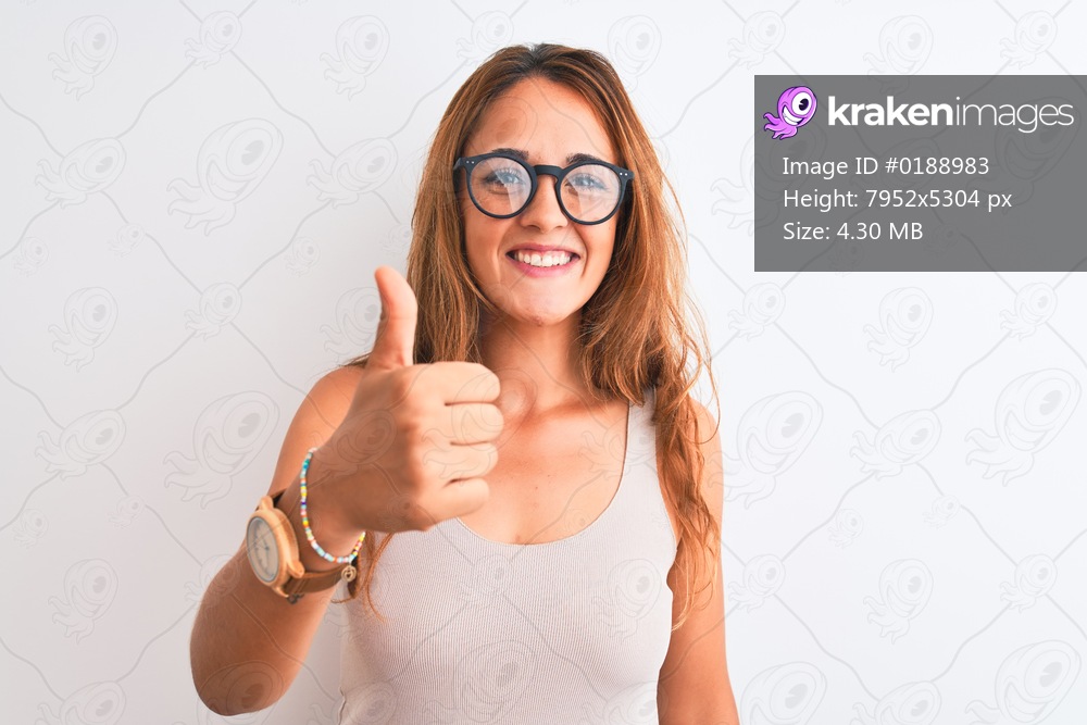 Young redhead woman wearing glasses standing over white isolated background doing happy thumbs up gesture with hand. Approving expression looking at the camera with showing success.