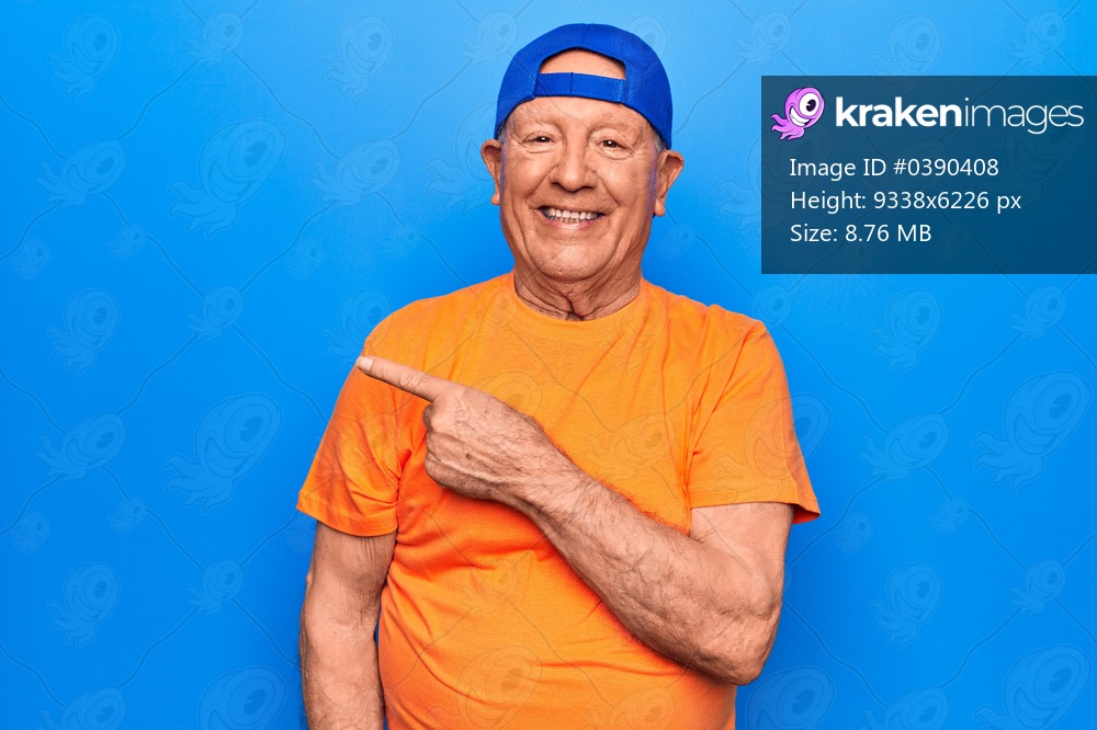Senior handsome grey-haired man wearing casual t-shirt and cap over blue background smiling cheerful pointing with hand and finger up to the side