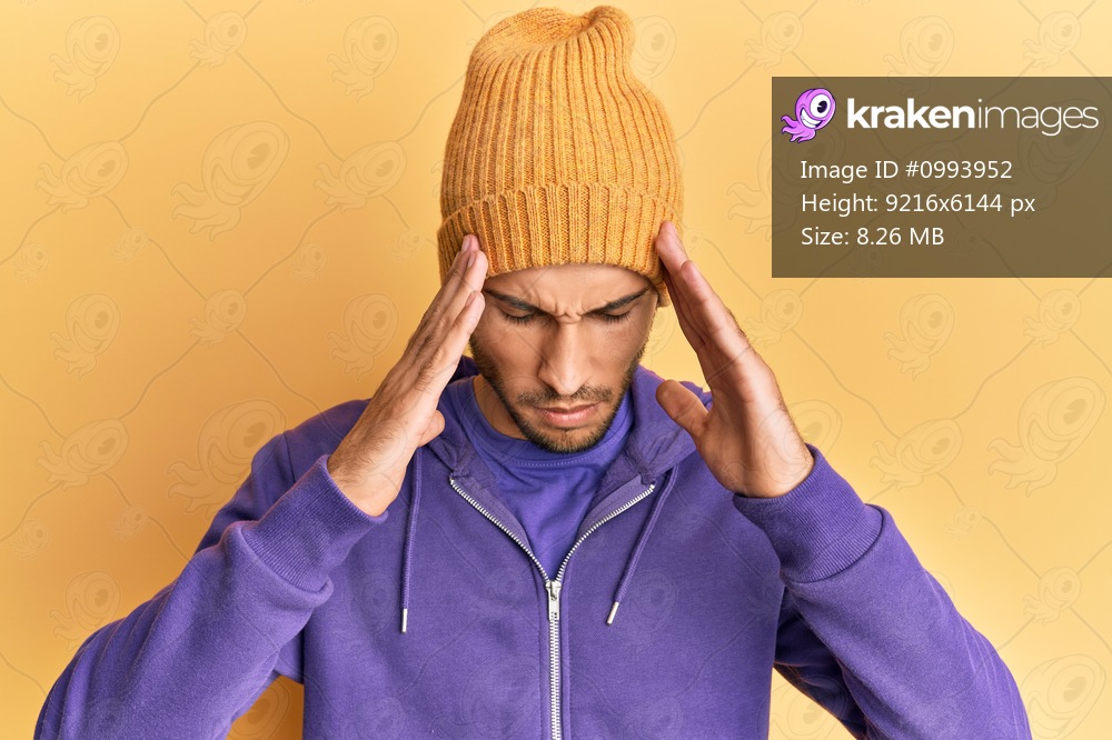 Young handsome man wearing wool winter hat with hand on head, headache because stress. suffering migraine. 