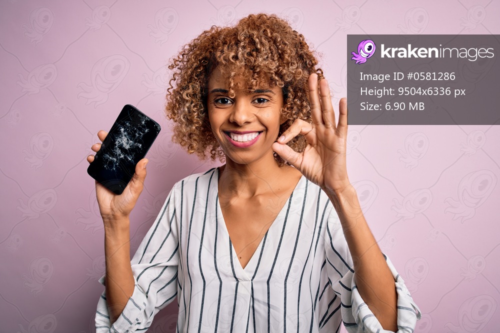 Young african american curly woman holding broken smartphone showing craked screen doing ok sign with fingers, excellent symbol